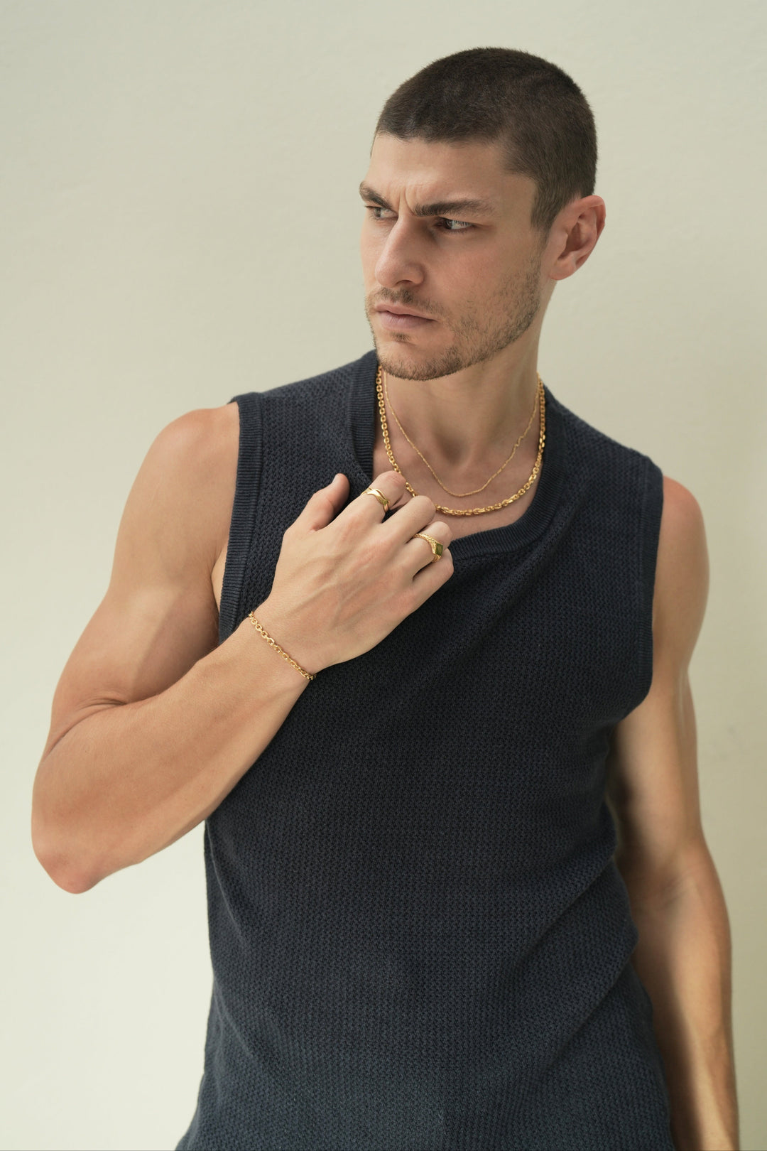 Founder of Nusands Corey Micari waist up in navy tank top looking to the right of camera, right hand grasping neckline of tank top, wearing 18k gold shackled bracelet, stacked wave rings and suns out ring. Around his neck is stacked shackled necklace and signature chain necklace