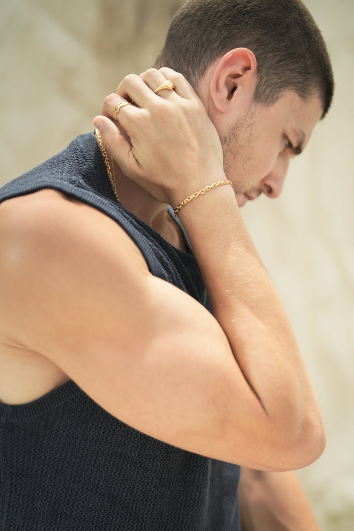 Founder of Nusands Corey Micari side profile with his right arm grasping the back of his neck, wearing matching 18k shackled bracelet and necklace that is stacked with signature chain necklace. On his write hand is stacked wave rings and a suns out ring.
