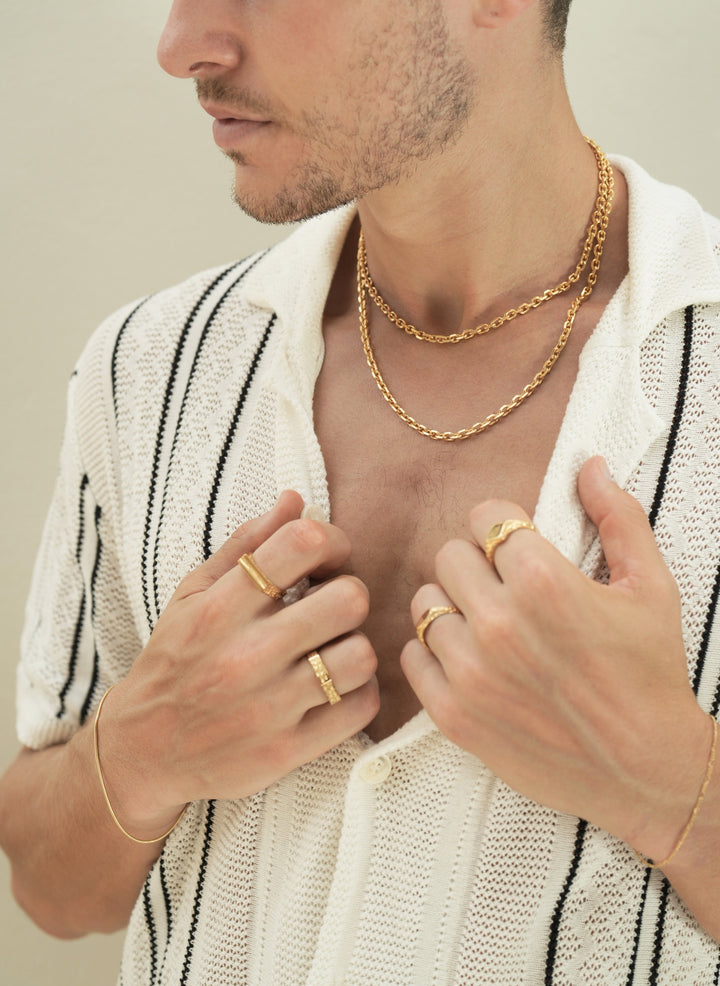 Founder of Nusands Corey Micari chest shot of open white shirt with black stripes unbuttoned to reveal stacked 45 and 55cm 18k gold shackled necklaces, looking away to the right, both hands grasping opening of shirt, roped in bracelet on right wrist, line in the and ring on index finger, six base on another. Left hand with mirrored signet on ring finger, suns out ring on index, signature chain bracelet on wrist.