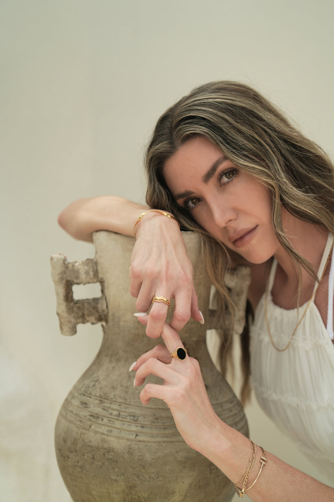 Founder of Nusands Emily Micari crouched over pot, head learning on right arm that is wearing 18K gold modest cuff and stacked wave rings on middle finger. slightly holding left hand that is wearing midnight onyx ring on that finger.