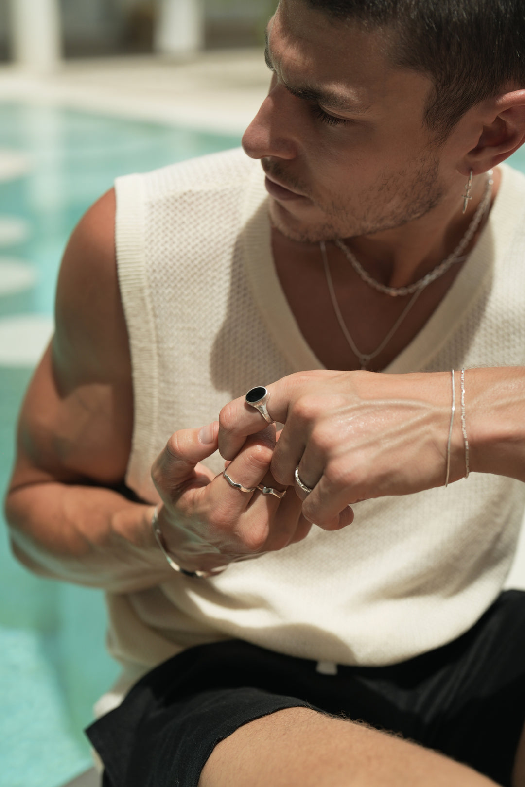Founder of Nusands Corey Micari, sitting, looking to the right of camera, wearing sterling silver hoop earring with sword charm, stacked shackled necklace with roped in necklace and lightning bolt charm. Both hands lightly clasping one another, modest cuff on right wrist, unstacked wave rings on middle and index fingers, mirrored signet ring on pink. Left hand has stacked roped in bracelet and signature chain bracelet, midnight onyx ring on index finger, stacker ring on ring finger.