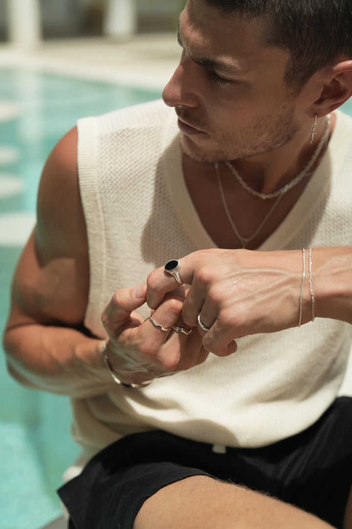 Founder of Nusands Corey Micari, sitting, looking to the right of camera, wearing sterling silver hoop earring with sword charm, stacked shackled necklace with roped in necklace and lightning bolt charm. Both hands lightly clasping one another, modest cuff on right wrist, unstacked wave rings on middle and index fingers, mirrored signet ring on pink. Left hand has stacked roped in bracelet and signature chain bracelet, midnight onyx ring on index finger, stacker ring on ring finger.