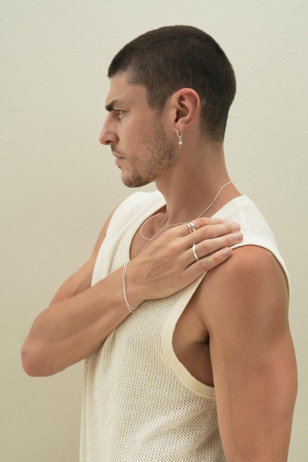 Founder of Nusands Corey Micari, side profile, right arm across body on left shoulder., wearing sterling silver hoop earring with lightning bolt charm, roped in necklace. Left hand has stacked roped in bracelet and signature chain bracelet, stacked wave rings on index finger, stacker ring on other finger.