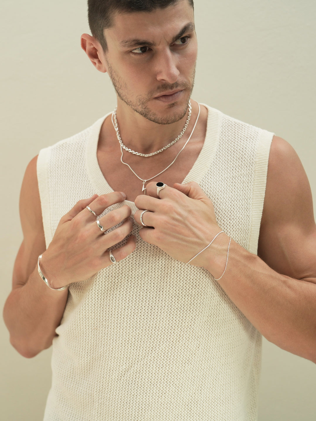 Founder of Nusands Corey Micari, chest up view of him looking to the left of camera, wearing sterling silver hoop earring with sword charm, stacked shackled necklace with roped in necklace and lightning bolt charm. Both hands graping neckline on white singlet, modest cuff on right wrist, unstacked wave rings on middle and index fingers, mirrored signet ring on pink. Left hand has stacked roped in bracelet and signature chain bracelet, midnight onyx ring on index finger, stacker ring on ring finger.
