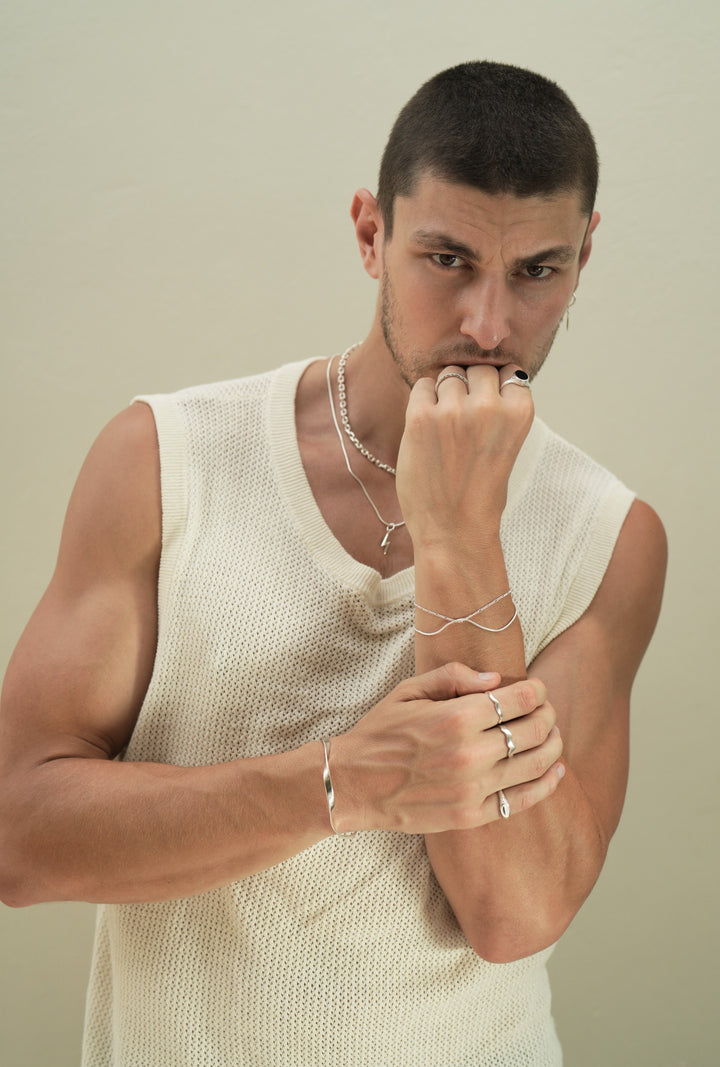 Founder of Nusands Corey Micari, chest up view of him looking to the to the camera, wearing sterling silver hoop earring with sword charm, stacked shackled necklace with roped in necklace and lightning bolt charm. left hand to chin, right hand grapsing left forearm, modest cuff on right wrist, unstacked wave rings on middle and index fingers, mirrored signet ring on pink. Left hand has stacked roped in bracelet and signature chain bracelet, midnight onyx ring on index finger, stacker ring on ring finger.
