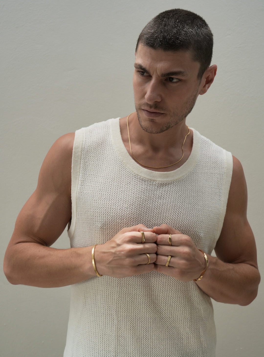 Founder of Nusands Corey Micari wearing 18k gold roped in necklace around neck, right hand with stirking cuff, line in the sand and stacker ring. Modest cuff on left wrist, mirrored signet and six base ring on hand fists together front profile, look away to right of camera, waist high view, white singlet on.