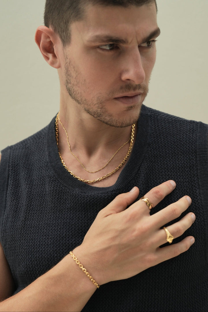 Founder of Nusands Corey Micari chest up front on view with him looking to the left of the camera wearing a blue singlet, right hand flat across his chest. Stacked signature chain and shackled 18k gold necklaces around his neck. Stacked wave rings on right hands index finger, suns out ring on another. Matching shackled bracelet around his wrist.
