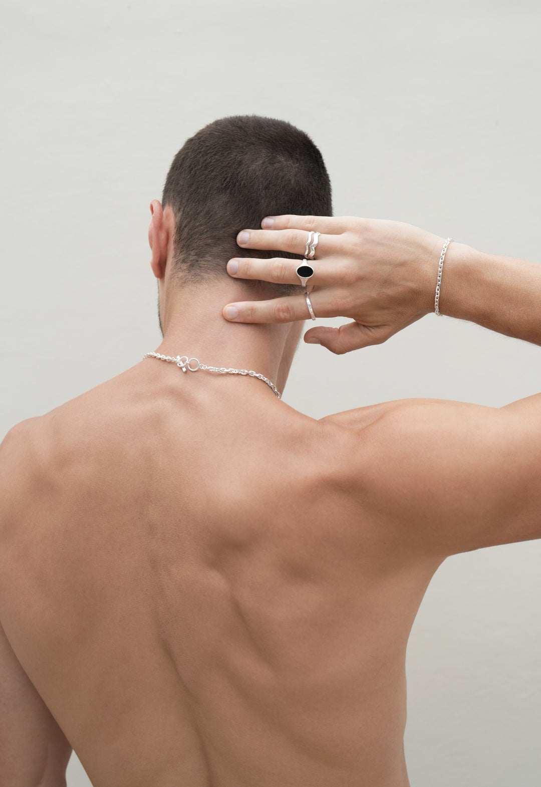 Founder of Nusands Corey Micari, waist up view of his back, right hand touching back of head, with sterling silver jewellery, stacked wave rings on a finger, midnight onyx ring on middle finger, stacker ring on index finger. Shackled bracelet on his right wrist to match the shackled bracelet around his neck.
