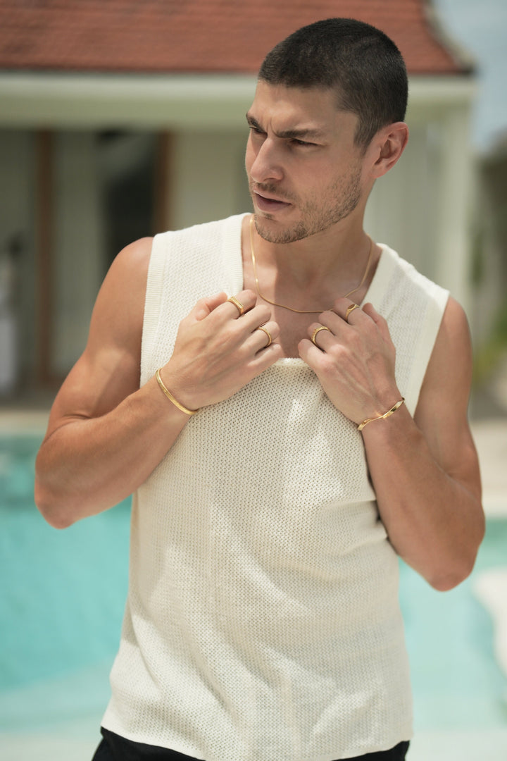 Founder of Nusands Corey Micari front on view looking away to the right both hands at the top of singlet grasping at neckline. 18k gold Roped in necklace is worn with stirking cuff on the right wrist and line in the sand and stacker rings on fingers. Left arm has modest cuff around wrist with six base and mirrored signet on fingers.