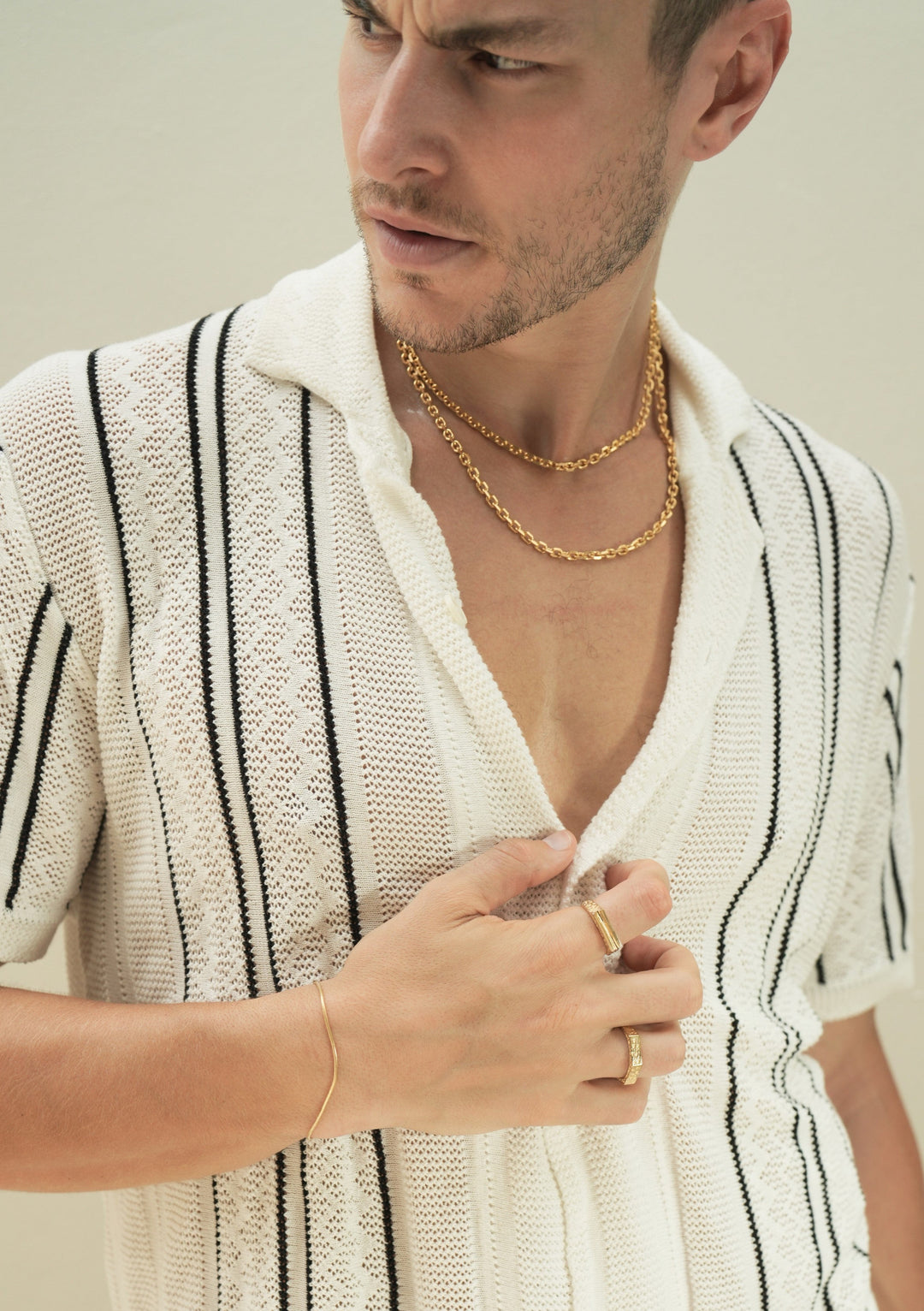 Founder of Nusands Corey Micari chest shot of open white shirt with black stripes unbuttoned to reveal stacked 45 and 55cm 18k gold shackled necklaces, looking away to the right, right hand grasping opening of shirt, roped in bracelet on right wrist, line in the and ring on index finger, six base on another.