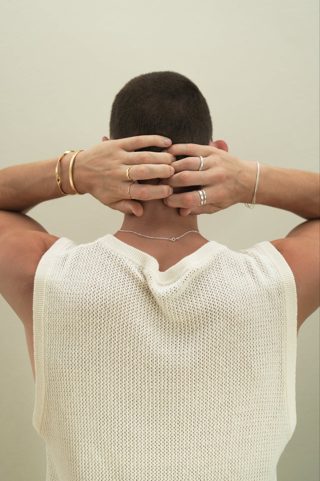 Founder of Nusands Corey Micari view of waist up back of body, hands lightly clenched behind his head, neck has sterling silver signature chain around it. Left wrist has stacked 18k gold modest and striking cuff, hand has unstacked wave rings across 2 fingers. Right hand has stacked sterling silver signature chain and roped in bracelets, stacked wave rings on ring finger, stacker ring on index.