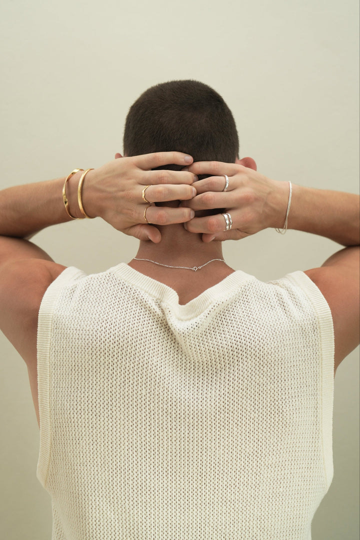 Founder of Nusands Corey Micari view of waist up back of body, hands lightly clenched behind his head, neck has sterling silver signature chain around it. Left wrist has stacked 18k gold modest and striking cuff, hand has unstacked wave rings across 2 fingers. Right hand has stacked sterling silver signature chain and roped in bracelets, stacked wave rings on ring finger, stacker ring on index.