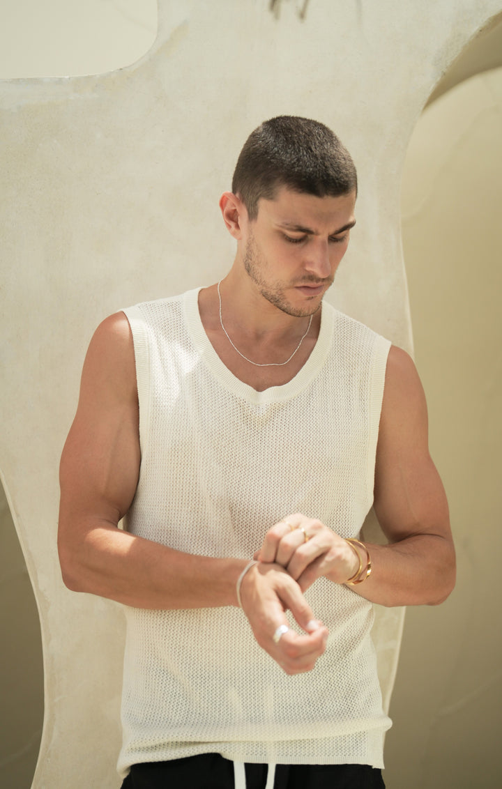 Founder of Nusands Corey Micari waist high front on view, he is looking down slightly to the right. Sterling Silver signature chain necklace around his neck, left hand adjusting shackled bracelet on his right wrist, that also has stacked wave rings on the right index finger. His left hand wears 18k gold unstacked wave rings across two fingers and modest cuff around his wrist.