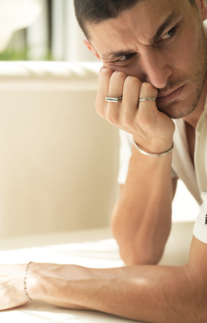 Founder of Nusands Corey Micari leaning over a table looking to the left of camera, right wrist propping head up, line in the sand sterling silver ring on middle finger, stacker ring on pinky, striking cuff on right wrist. Shacked bracelet on left wrist.