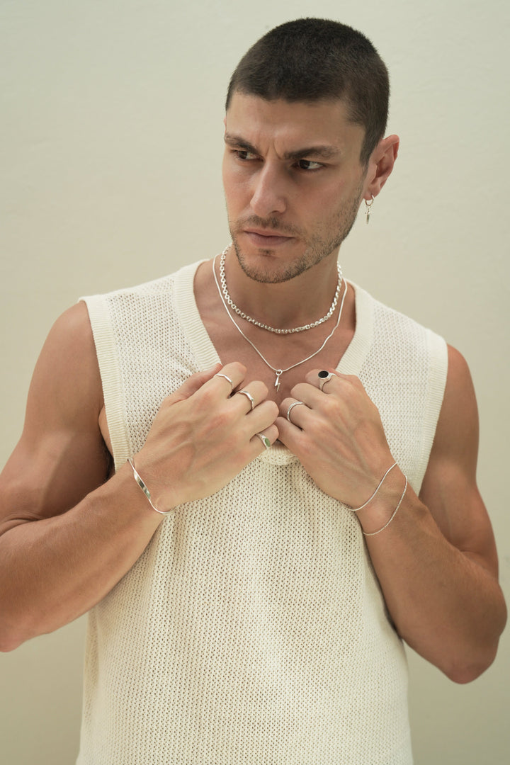 Founder of Nusands Corey Micari front on view, looking to the right of camera, both hands grasping neckline of white singlet. Stacked sterling silver shackled and signature chain necklaces with lightning both charm. Right arm is wearing the modest cuff and unstacked wave rings with mirrored signet ring. Left had wearing stacking roped in and signature chain bracelets, stacker and midnight onyx rings.