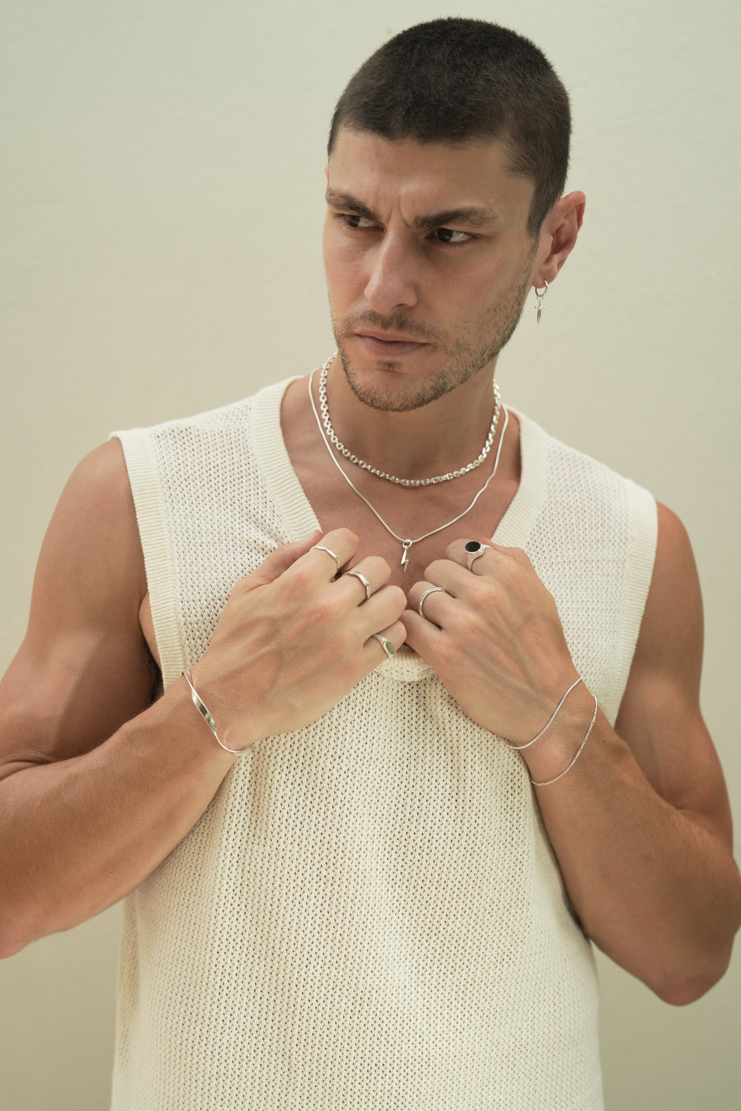 Founder of Nusands Corey Micari front on view, looking to the right of camera, both hands grasping neckline of white singlet. Stacked sterling silver shackled and signature chain necklaces with lightning both charm. Right arm is wearing the modest cuff and unstacked wave rings with mirrored signet ring. Left had wearing stacking roped in and signature chain bracelets, stacker and midnight onyx rings.