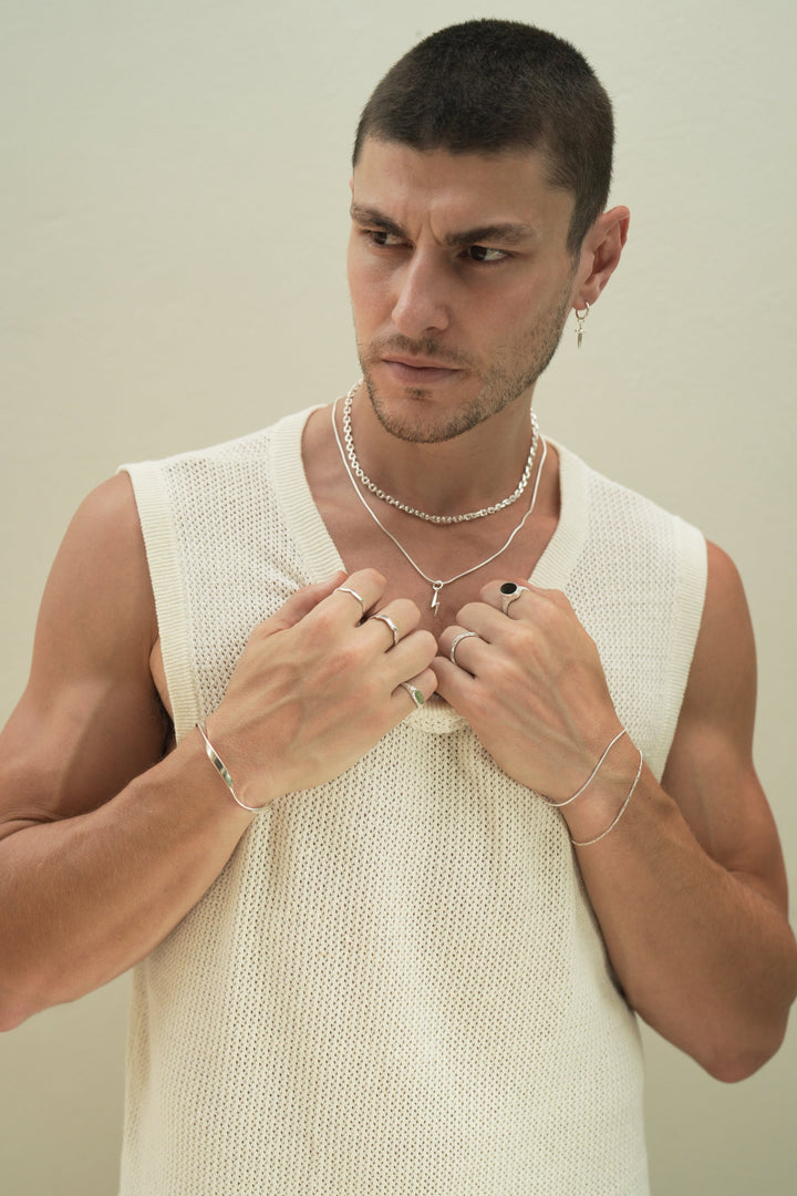 Founder of Nusands Corey Micari front on view, looking to the right of camera, both hands grasping neckline of white singlet. Stacked sterling silver shackled and signature chain necklaces with lightning both charm. Right arm is wearing the modest cuff and unstacked wave rings with mirrored signet ring. Left had wearing stacking roped in and signature chain bracelets, stacker and midnight onyx rings.