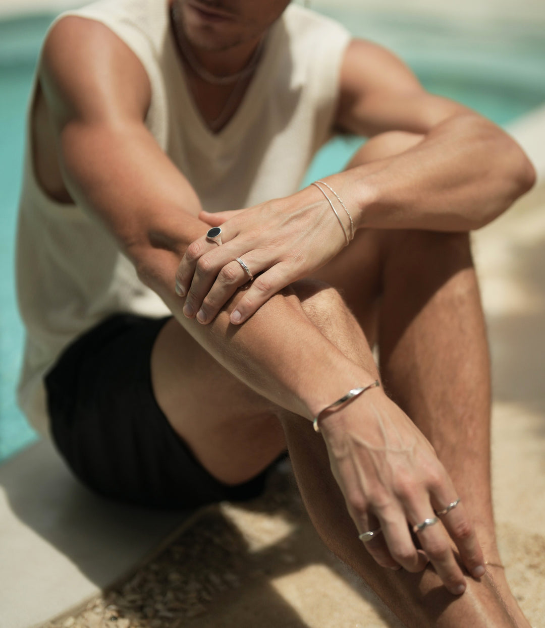 Founder of Nusands Corey Micari sitting by the pool right arm straight wearing sterling silver modest cuff on wrist, mirrored signet ring on pinky, unstacked wave ring on middle and index fingers. Left arm is grasping right forearm, stacker ring on ring finger, midnight onyx ring on index finger. Stacked roped in and signature chain bracelets on wrist.