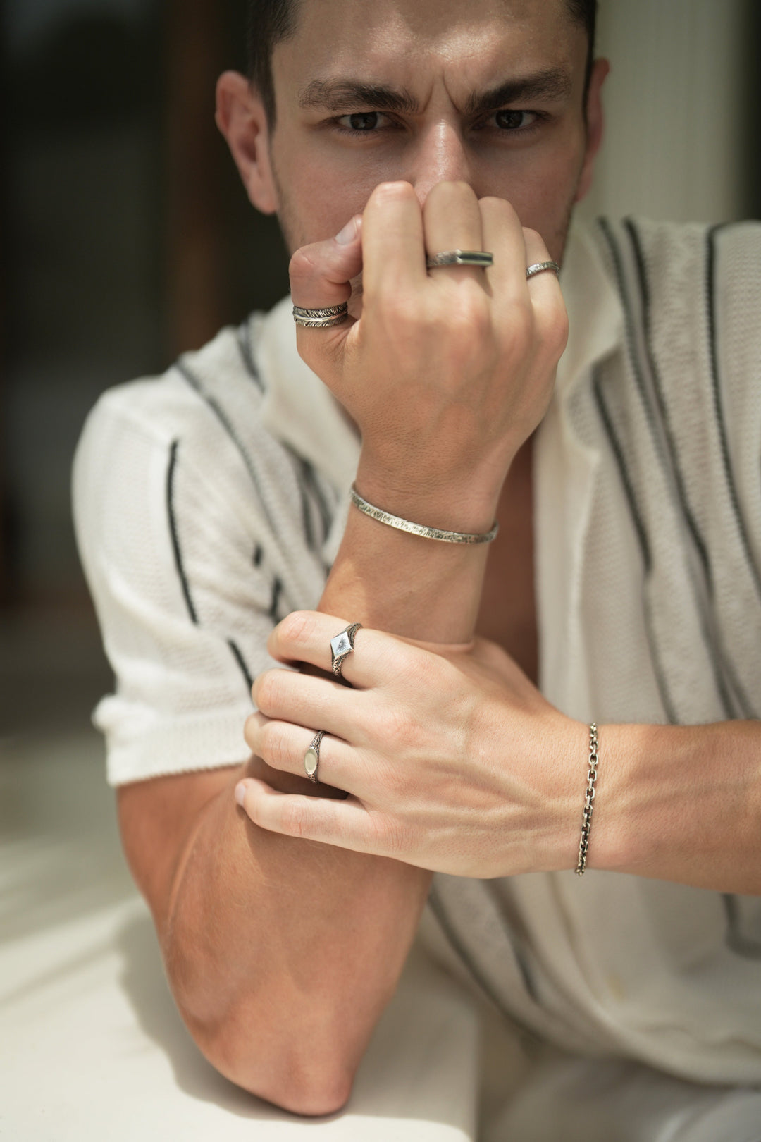 Founder of Nusands Corey Micari front on view staring down the camera, leaning on a table right fist infront of bottom half of his face wearing sterling silver striknig cuff, blade, line in the sand and stacker rings. Left hand grasping right forearm wearing shackled bracelet, suns out and mirrored signet rings.