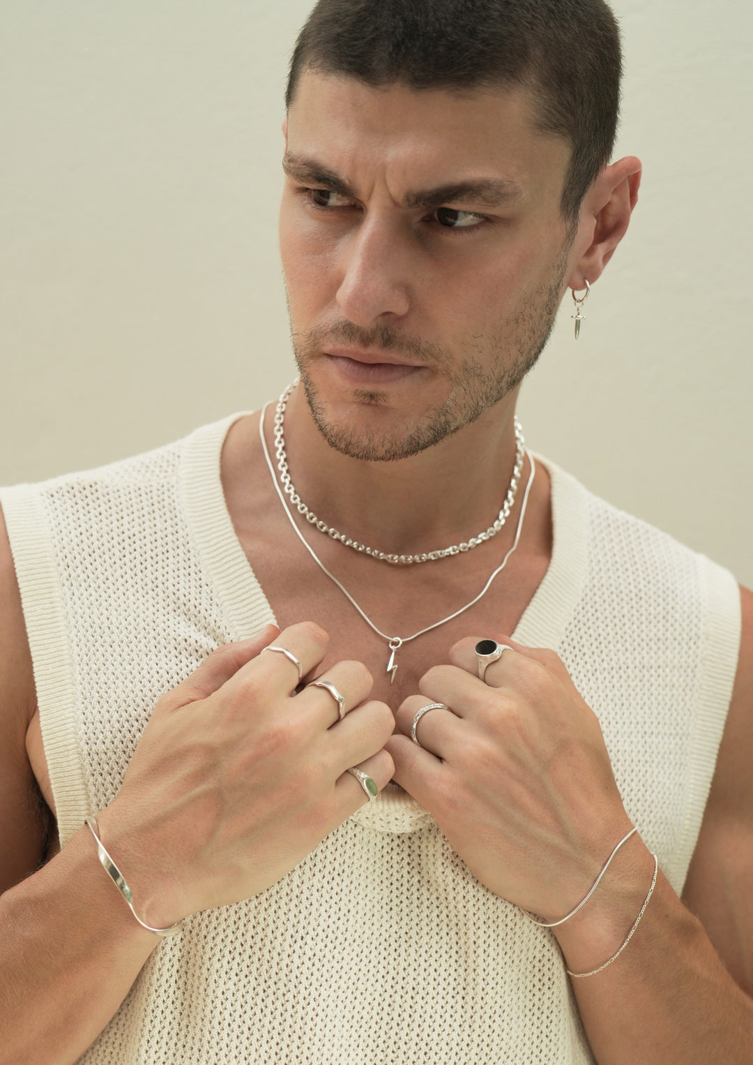 Founder of Nusands Corey Micari, chest up view of him looking to the right of camera, wearing sterling silver hoop earring with sword charm, stacked shackled necklace with roped in necklace and lightning bolt charm. Both hands grabbing neckline on white singlet, modest cuff on right wrist, unstacked wave rings on middle and index fingers, mirrored signet ring on pinky. Left hand has stacked roped in bracelet and signature chain bracelet, midnight onyx ring on index finger, stacker ring on ring finger.