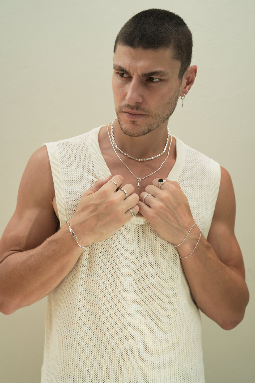 Founder of Nusands Corey Micari,  waist up view of him looking to the right of camera, wearing sterling silver hoop earring with sword charm, stacked shackled necklace with roped in necklace and lightning bolt charm. Both hands grabbing neckline on white singlet, modest cuff on right wrist, unstacked wave rings on middle and index fingers, mirrored signet ring on pinky. Left hand has stacked roped in bracelet and signature chain bracelet, midnight onyx ring on index finger, stacker ring on ring finger.