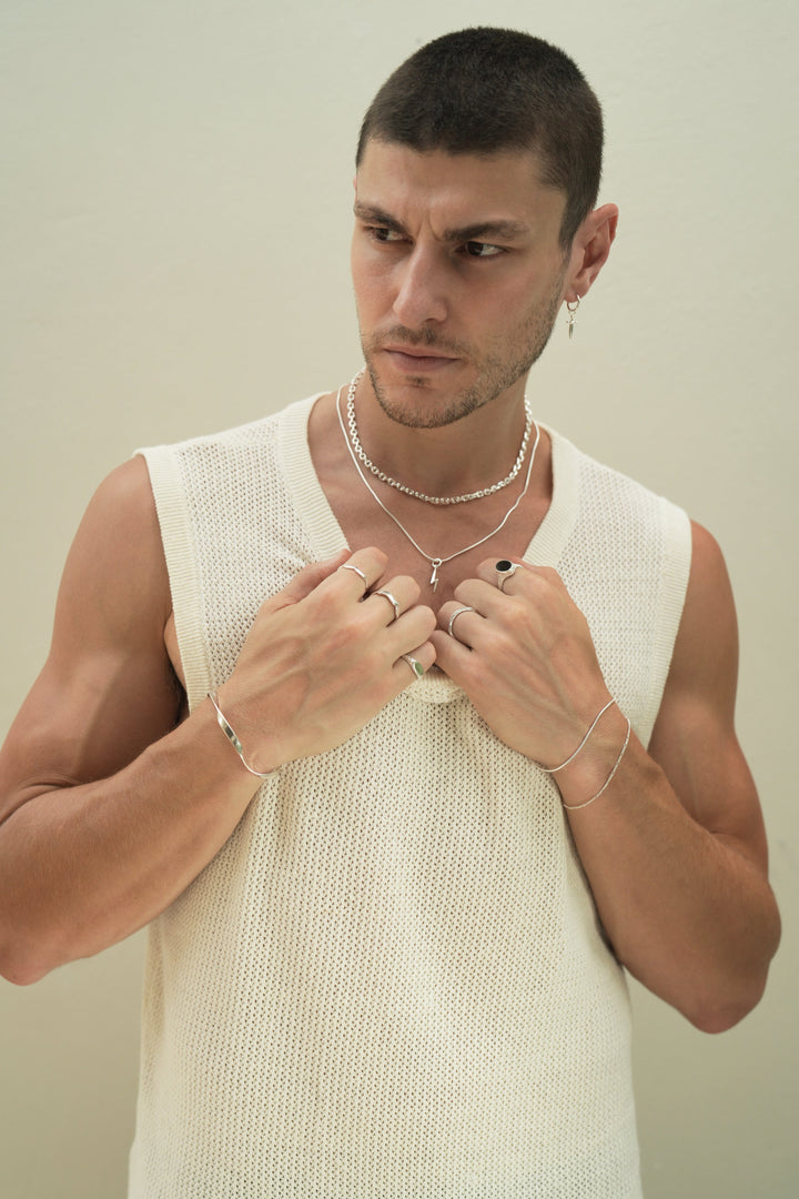 Founder of Nusands Corey Micari,  waist up view of him looking to the right of camera, wearing sterling silver hoop earring with sword charm, stacked shackled necklace with roped in necklace and lightning bolt charm. Both hands grabbing neckline on white singlet, modest cuff on right wrist, unstacked wave rings on middle and index fingers, mirrored signet ring on pinky. Left hand has stacked roped in bracelet and signature chain bracelet, midnight onyx ring on index finger, stacker ring on ring finger.