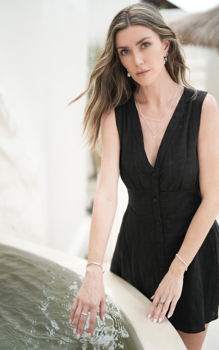 Founder of Nusands Emily Micari front profile standing in front of water, black dress left hand on edge of water wearing sterling silver shackled bracelet and line in the sand ring, right hand on top of water wearing modest cuff and wave rings, signature chain necklace with lightning bolt charm and roped in necklace around her neck, staring at camera