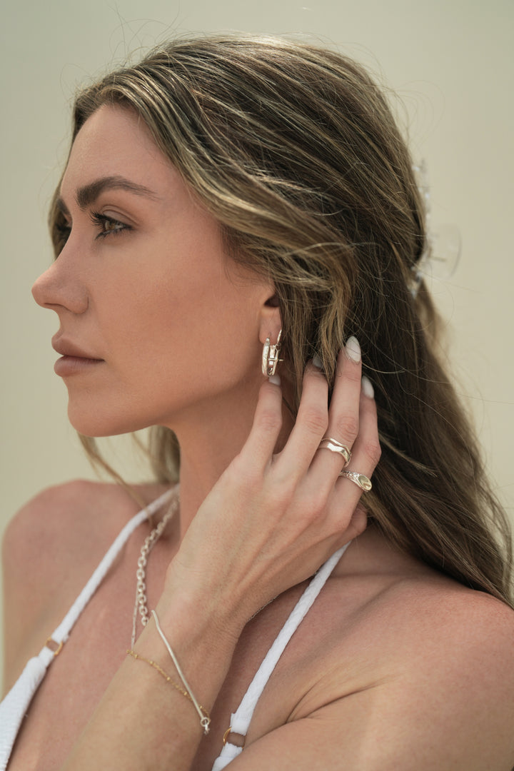 Founder of Nusands Emily Micari side profile of head. Left hand grasping her neck, stacked sterling silver wave rings on middle finger, mirrored signet on index. Stacked wave and cross earrings in left ear. stacked shackled and signature chain necklaces around neck.