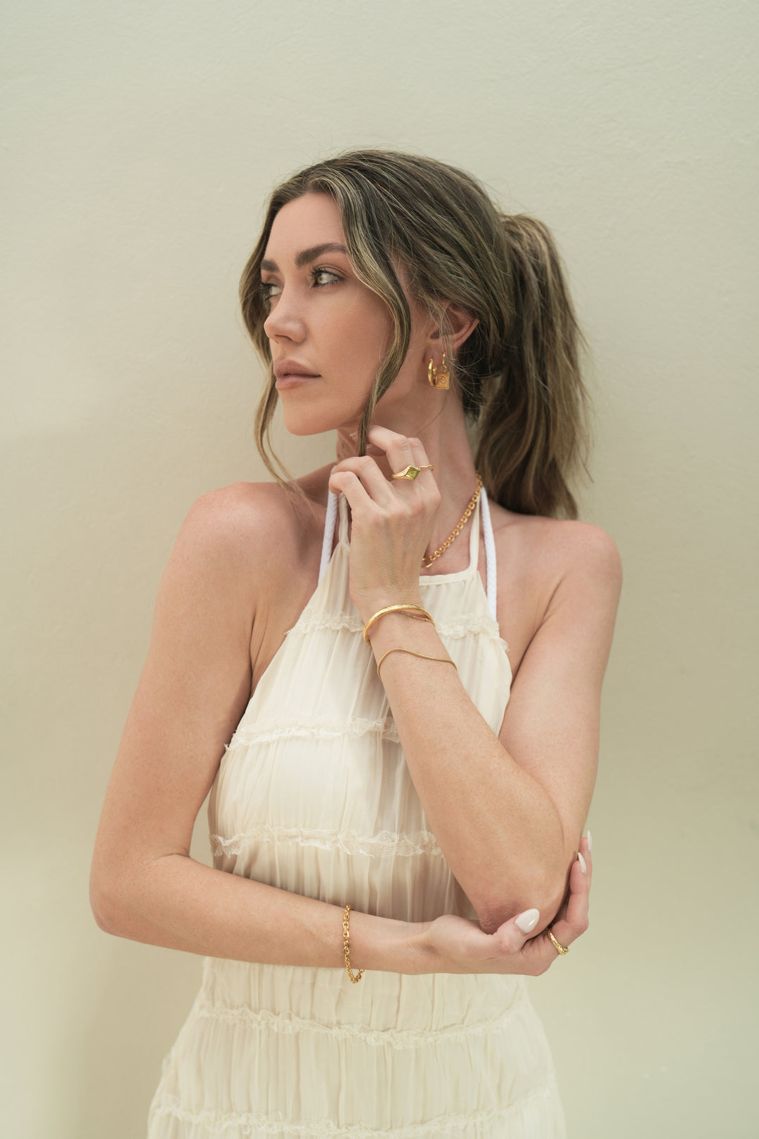 Founder of Nusands Emily Micari, waist up view, looking away to the right, white dress. Right hand underneath left elbow across her waist. 18k gold shackled bracelet on wrist, stacker ring on index finger. striking cuff and roped in bracelet stacked on left wrist, her hand under her chin, suns out ring on middle finger, hoop earrings with evil eye charm in left ear.