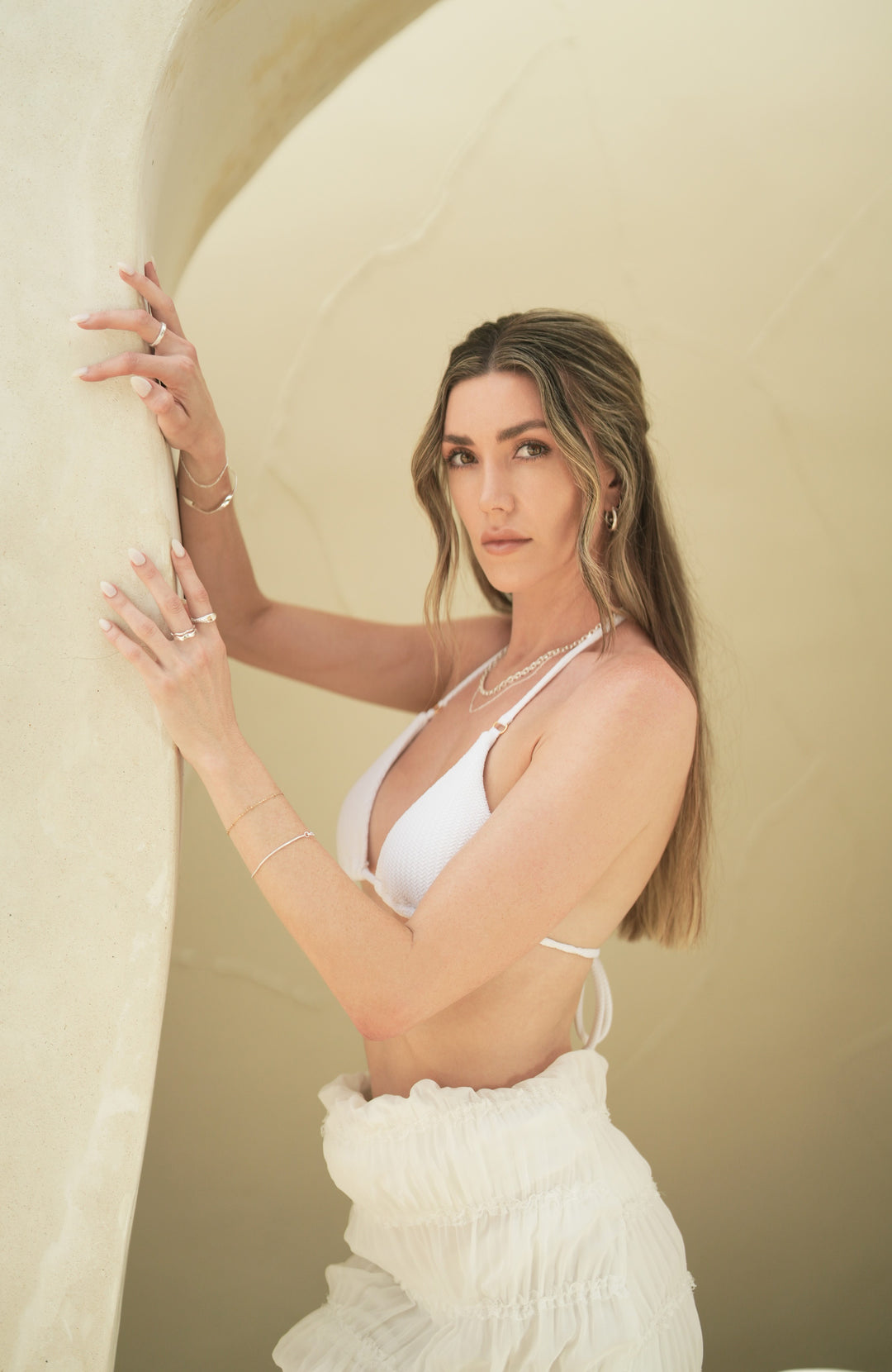 Founder of Nusands Emily Micari, both hands on wall looking to camera white bikini top. Stacked sterling silver shackled and signature chain necklaces. Left wrist has roped in bracelet, stacked wave ring on middle finger, mirrored signet on index finger. Right hands has stacker ring on middle finger, modest cuff on wrist.