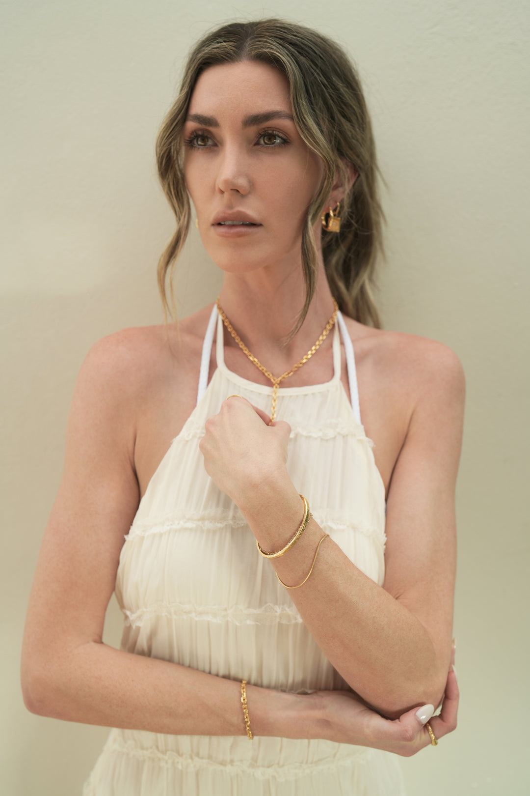 Founder of Nusands Emily Micari waist high front on shot, her looking to the right of camera, right hand grasping left elbow infront of her body. 18K gold evil eye earrings in her left ear, shackled necklace around her neck being touched by her left hand matching her shackled bracelet on her right wrist. mirrored signet and stacker rings on fingers. Her left wrist has stacked striking cuff with signature chain bracelet, suns out and thinner wave rings on her fingers.