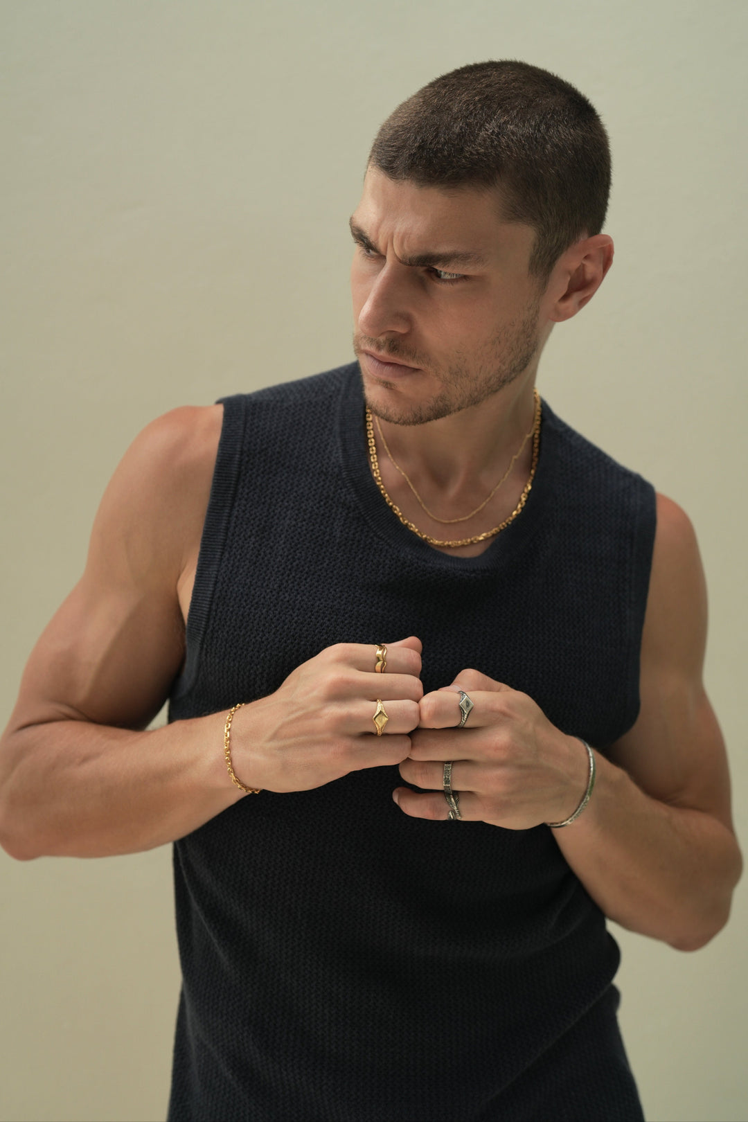 Founder of Nusands Corey Micari waist up shot staring to the right of camera wearing navy singlet, both hands meeting infront of chest in fists. Wearing 18K Gold stacked signature chain and shackled necklaces around neck, shackled bracelet on right wrist with suns out ring on a finger, stacked wave rings on another. His left arm is all sterling silver jewellery, stirking cuff on wrist, suns out, stacker and blade rings on hand.