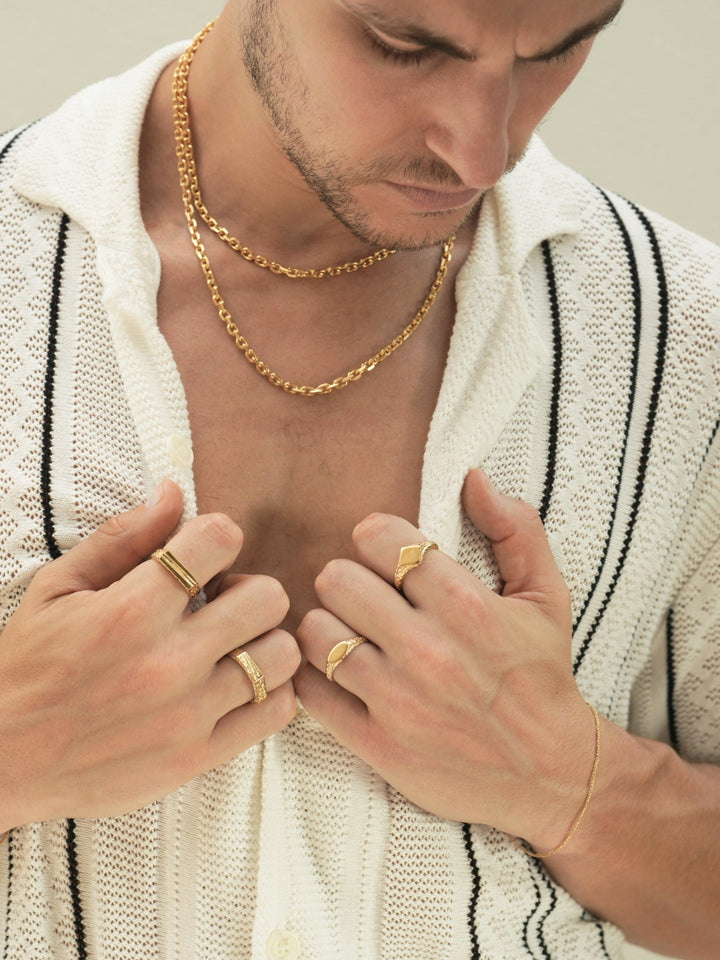 Founder of Nusands Corey Micari close up front chest view looking down, hands grasping inside of half unbuttoned white and black striped shirt. Stacked 45 and 55cm 18k gold shackled necklaces around his neck. Signature chain bracelet on left wrist, suns out and mirrored signet on separate fingers. Right hand has line in the sand and six base rings on separate fingers.