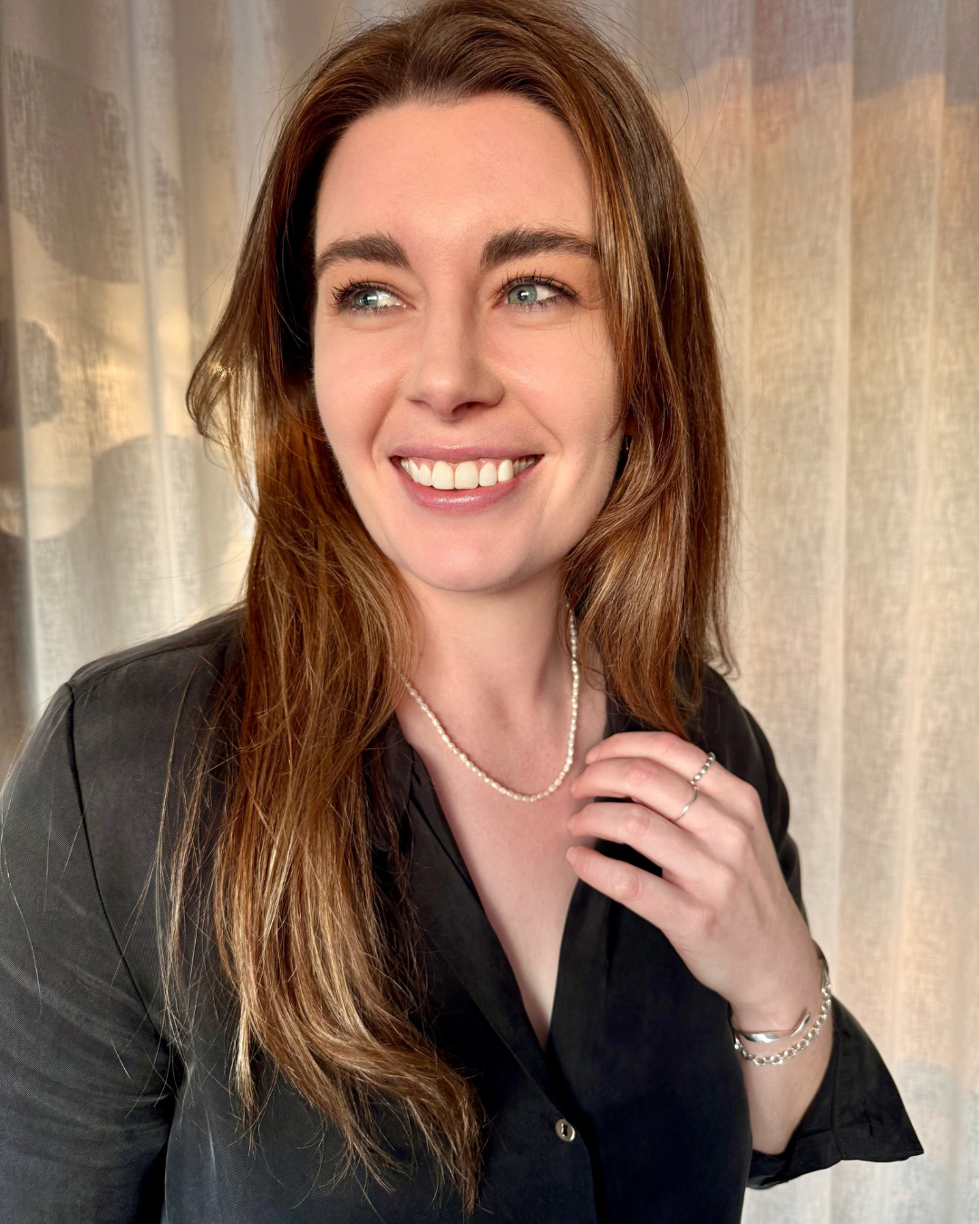 Woman with long brown hair wearing a black shirt and pearl necklace against a neutral background.
