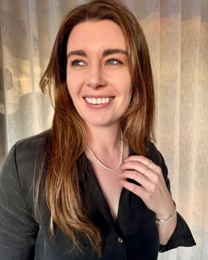Woman with long brown hair wearing a black shirt and pearl necklace against a neutral background.