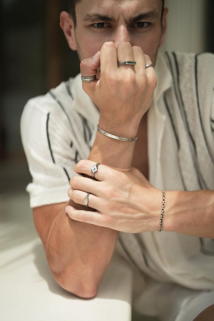 Founder of Nusands Corey Micari learning on a bench right fist infront of his face wearing sterling silver blade ring on his thumb, line in the sand ring on his middle finger and stacker ring on his pinky and striking cuff on his wrist. His left hand is grasping his right forearm with suns out and mirrored signet rings being worn and shackled bracelet on his wrist. staring directly at camera.