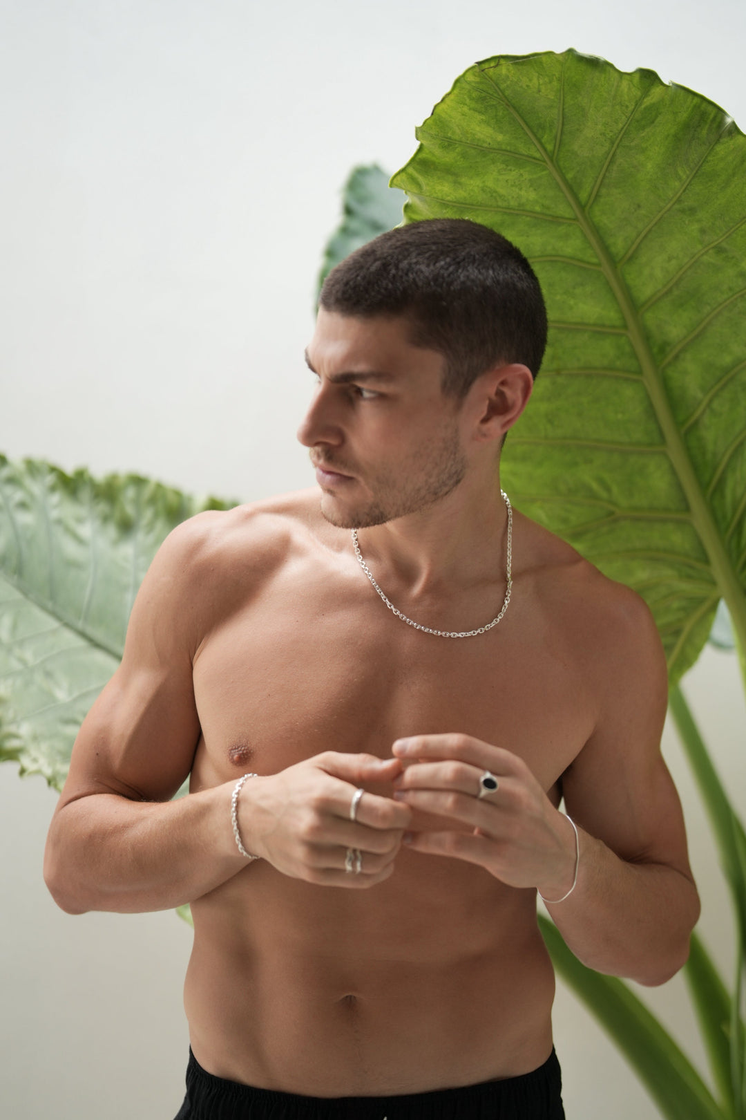 Founder of Nusands Corey Micari waist up front on view, shirtless looking to the right of camera. Sterling silve necklace around his neck, matched with his shackled bracelet on his right wrist. Both arms are half bent lightly touching fingers infront of his chest. Stacked wave rings on one of his fingers, stacker on another for his right hand. Left hand has the midnight onyx ring on his middle finger, roped in bracelet around his wrist.