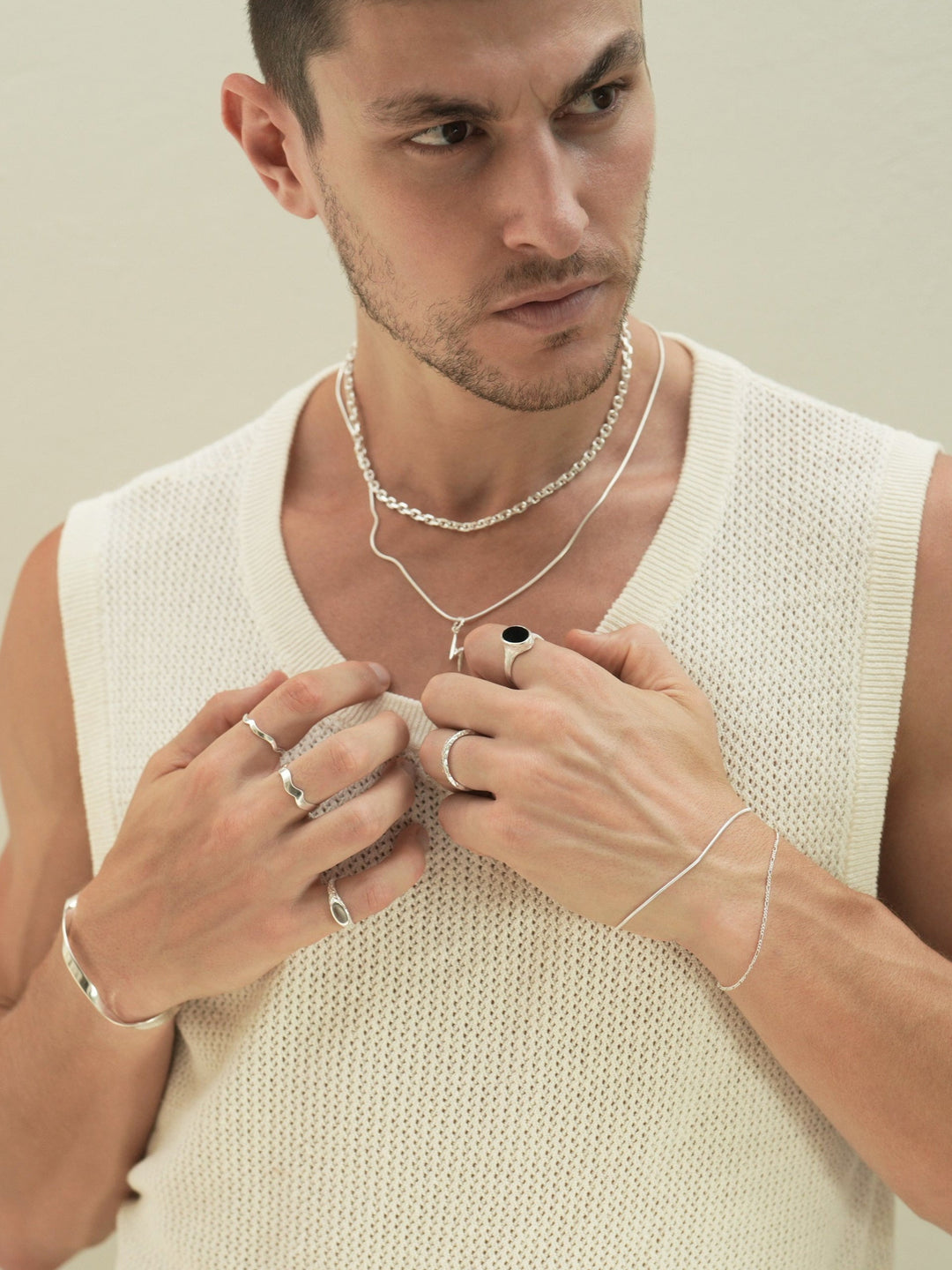 Founder of Nusands Corey Micari close front on chest looking to the left of camera wearing stacked sterling silver shackled necklace with roped in chain and cross charm. Hands lightly touching neckline of white singlet right hand slight higher wearing modest cuff, mirrored signet and wave rings on separate fingers. Left hand wearing stacked roped in and signature chain bracelets to match the necklaces, midnight onyx and stacker rings.