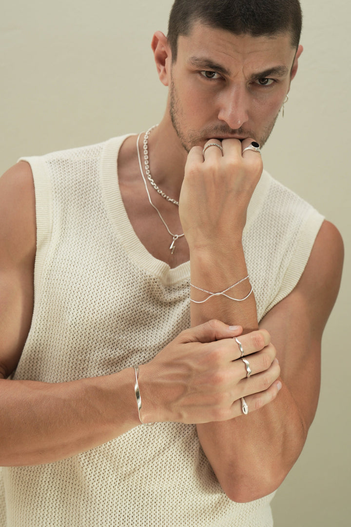 Founder of Nusands Corey Micari close front on chest looking straight at camera wearing stacked sterling silver shackled necklace with roped in chain and cross charm. left arm bent up, hand in a fist covering mouth, right arm lightly clasp left forearm wearing modest cuff, mirrored signet and wave rings on separate fingers. Left hand wearing stacked roped in and signature chain bracelets to match the necklaces, midnight onyx and stacker rings.