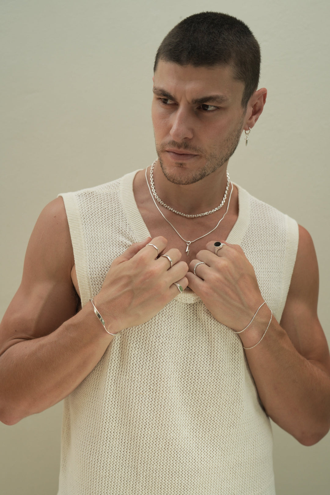 Founder of Nusands Corey Micari waist up front on view wearing a white singlet looking to the right of camera, both arms bent, hands grasping neckline of singlet. Sterling Silver modest cuff on right wrist, wave and mirrored signet rings worn on separate fingers, roped in and signature chain bracelets stacked on left wrist, stacker and midnight onyx rings on hand. Shackled and roped in necklaces stacked around neck with lightning bolt charm.