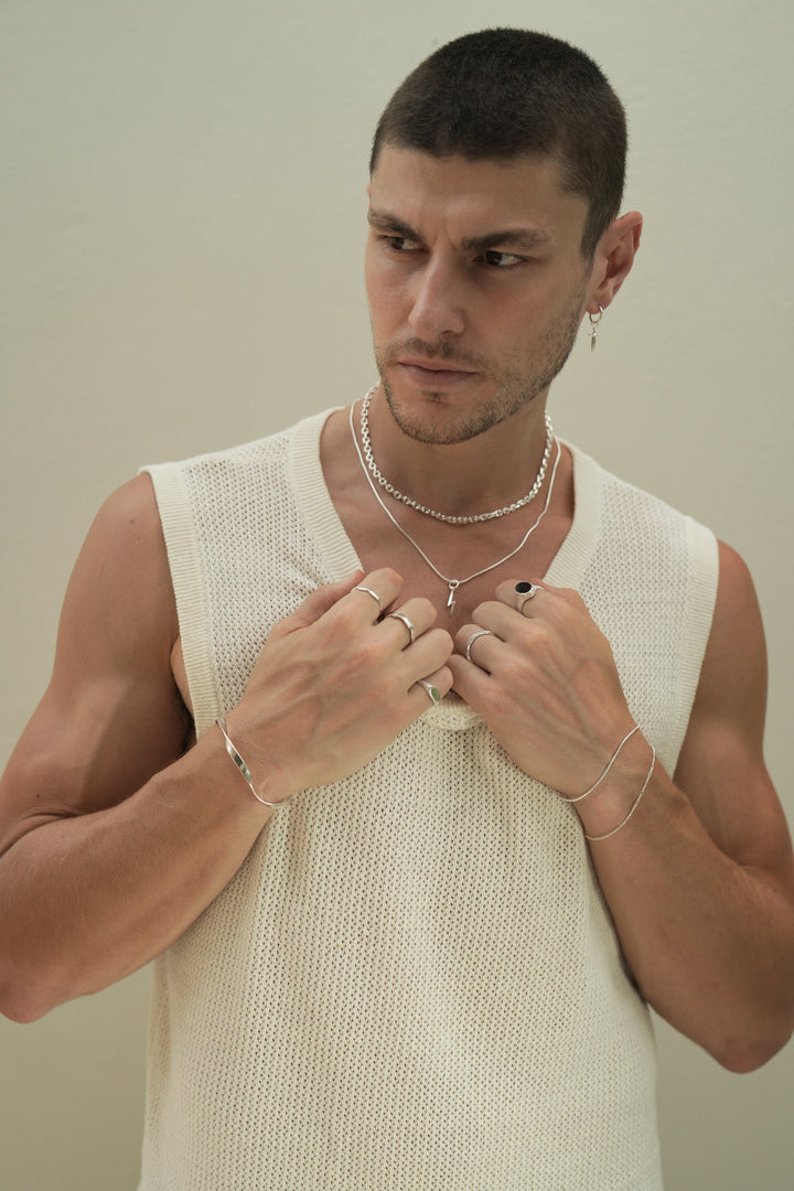 Founder of Nusands Corey Micari waist up front on view wearing a white singlet looking to the right of camera, both arms bent, hands grasping neckline of singlet. Sterling Silver modest cuff on right wrist, wave and mirrored signet rings worn on separate fingers, roped in and signature chain bracelets stacked on left wrist, stacker and midnight onyx rings on hand. Shackled and roped in necklaces stacked around neck with lightning bolt charm.