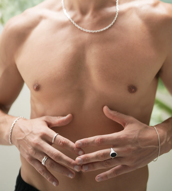 Founder of Nusands Corey Micari chest view, shirtless. Sterling silve necklace around his neck, matched with his shackled bracelet on his right wrist. Both arms are half bent lightly touching fingers infront of his chest. Stacked wave rings on one of his fingers, stacker on another for his right hand. Left hand has the midnight onyx ring on his middle finger, roped in bracelet around his wrist.