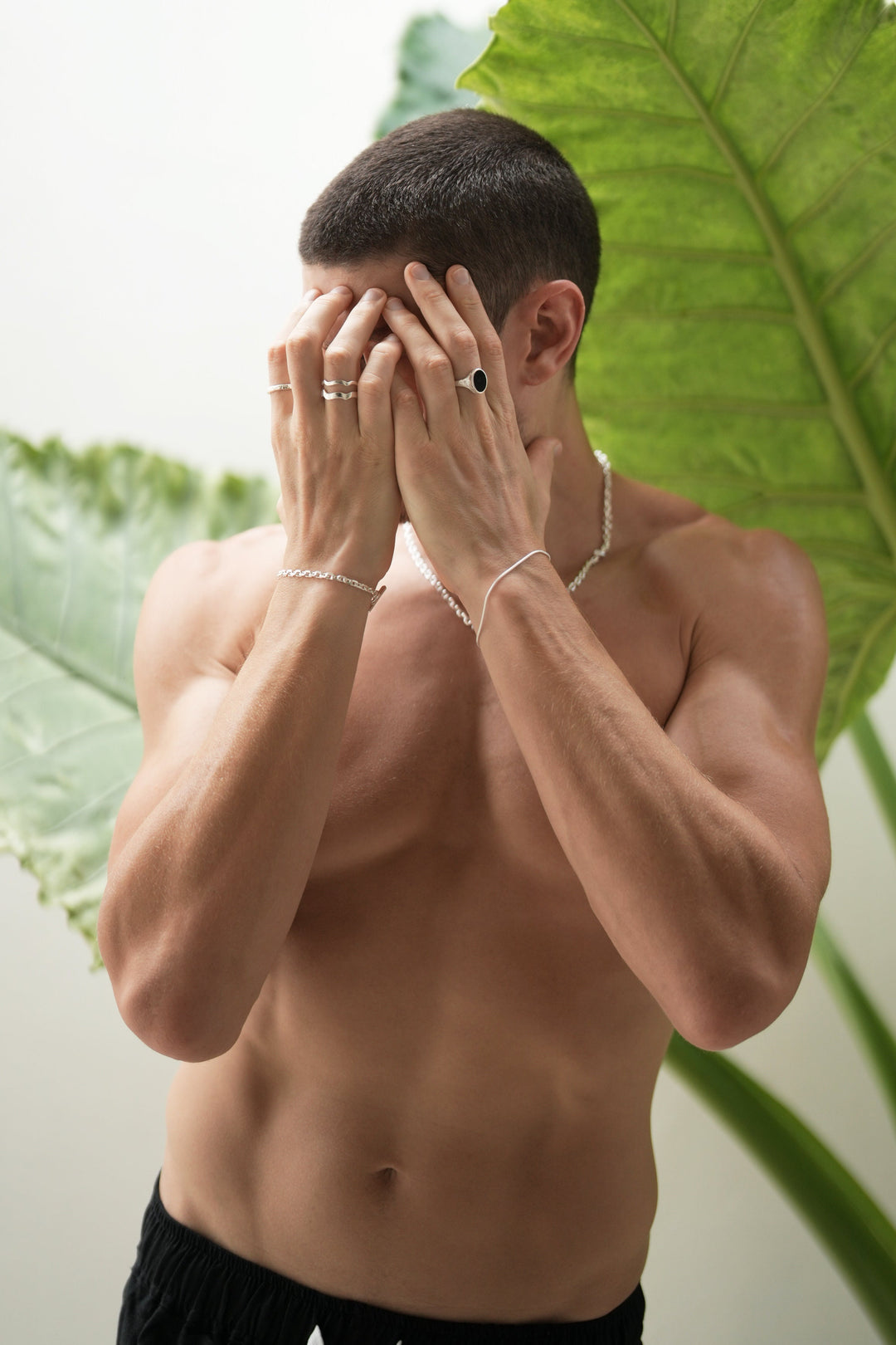 Founder Corey Micari waist up front on view, shirtless with both hands covering his face. Left wrist has Shiny sterling silver roped in bracelet and midnight onyx ring on hand. Right wrist is matching shackled bracelet with necklace, stacked wave rings and mirrored signet rings on hand.