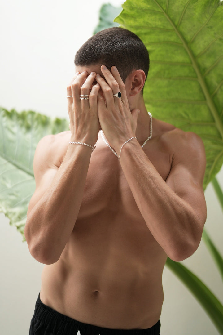 Founder Corey Micari waist up front on view, shirtless with both hands covering his face. Left wrist has Shiny sterling silver roped in bracelet and midnight onyx ring on hand. Right wrist is matching shackled bracelet with necklace, stacked wave rings and mirrored signet rings on hand.
