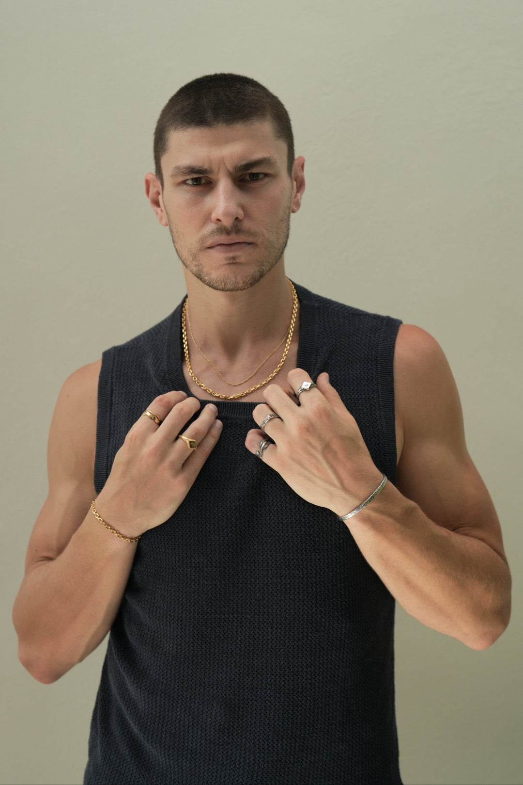 Founder of Nusands Corey Micari waist up shot staring at camera wearing navy singlet, both hands grasping neckling of the singlet. Wearing 18K Gold stacked signature chain and shackled necklaces around neck, shackled bracelet on right wrist with suns out ring on a finger, stacked wave rings on another. His left arm is all sterling silver jewellery, stirking cuff on wrist, suns out, stacker and blade rings on hand.