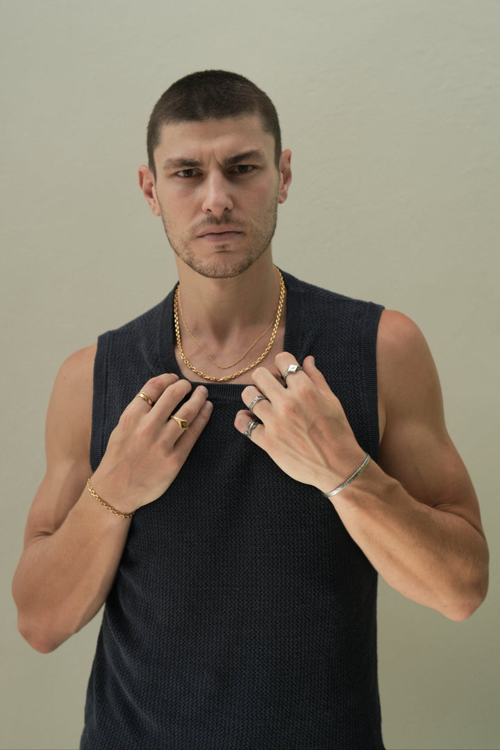 Founder of Nusands Corey Micari waist up shot staring at camera wearing navy singlet, both hands grasping neckling of the singlet. Wearing 18K Gold stacked signature chain and shackled necklaces around neck, shackled bracelet on right wrist with suns out ring on a finger, stacked wave rings on another. His left arm is all sterling silver jewellery, stirking cuff on wrist, suns out, stacker and blade rings on hand.