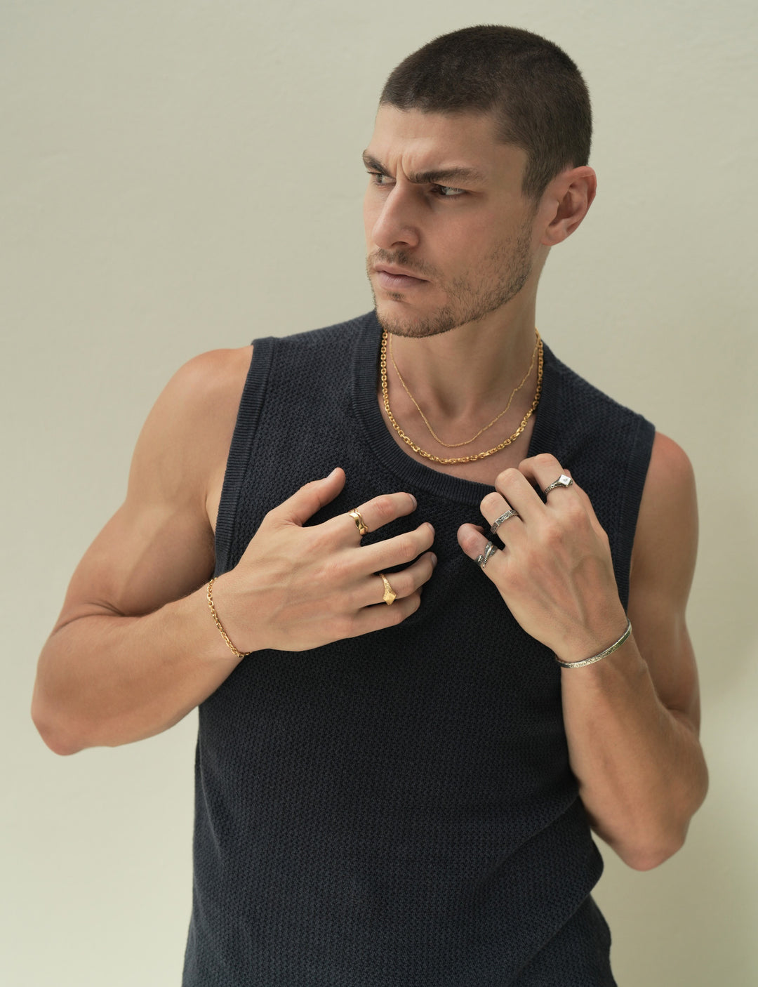Founder of Nusands Corey Micari waist up shot staring to the right of camera wearing navy singlet, both hands grasping neckling of the singlet. Wearing 18K Gold stacked signature chain and shackled necklaces around neck, shackled bracelet on right wrist with suns out ring on a finger, stacked wave rings on another. His left arm is all sterling silver jewellery, stirking cuff on wrist, suns out, stacker and blade rings on hand.