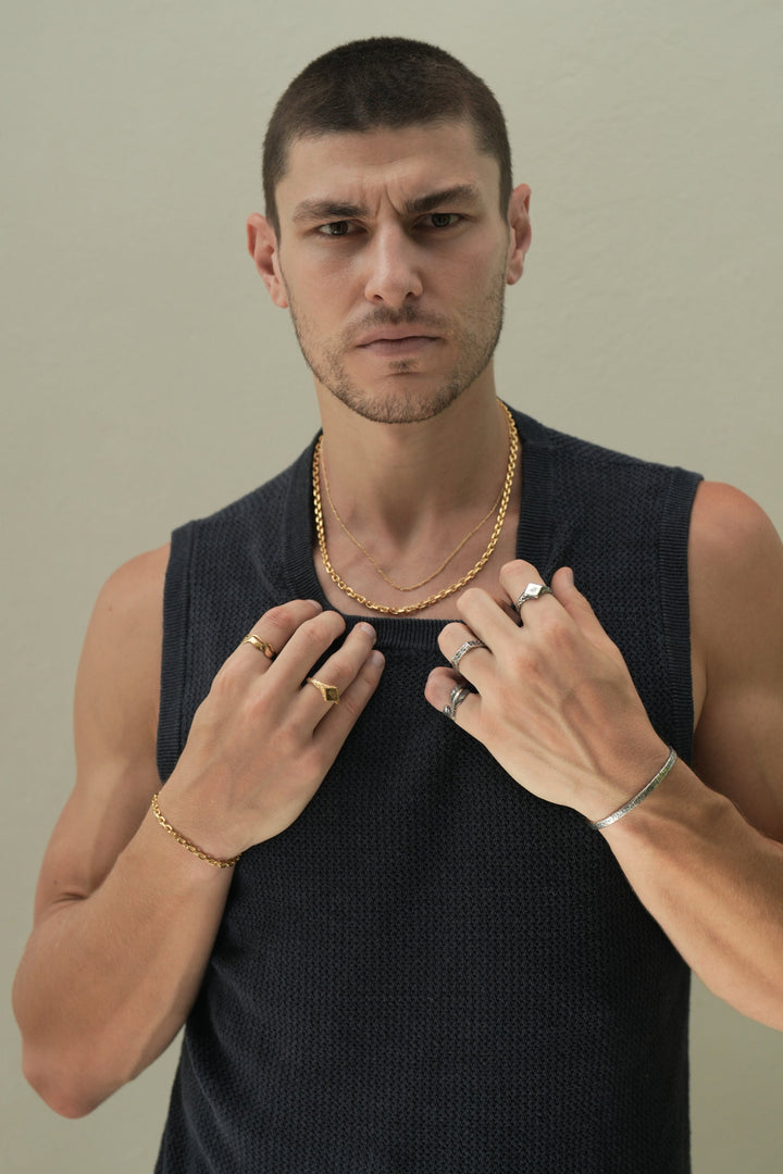 Founder of Nusands Corey Micari waist up shot staring at camera wearing navy singlet, both hands grasping neckling of the singlet. Wearing 18K Gold stacked signature chain and shackled necklaces around neck, shackled bracelet on right wrist with suns out ring on a finger, stacked wave rings on another. His left arm is all sterling silver jewellery, stirking cuff on wrist, suns out, stacker and blade rings on hand.