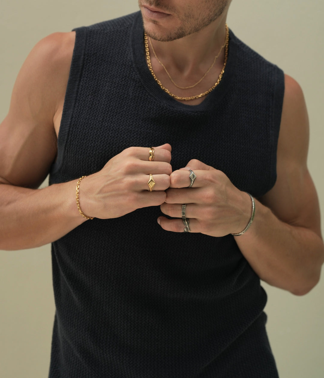 Founder of Nusands Corey Micari waist up shot staring to the right of camera wearing navy singlet, both hands meeting infront of chest in fists. Wearing 18K Gold stacked signature chain and shackled necklaces around neck, shackled bracelet on right wrist with suns out ring on a finger, stacked wave rings on another. His left arm is all sterling silver jewellery, stirking cuff on wrist, suns out, stacker and blade rings on hand.