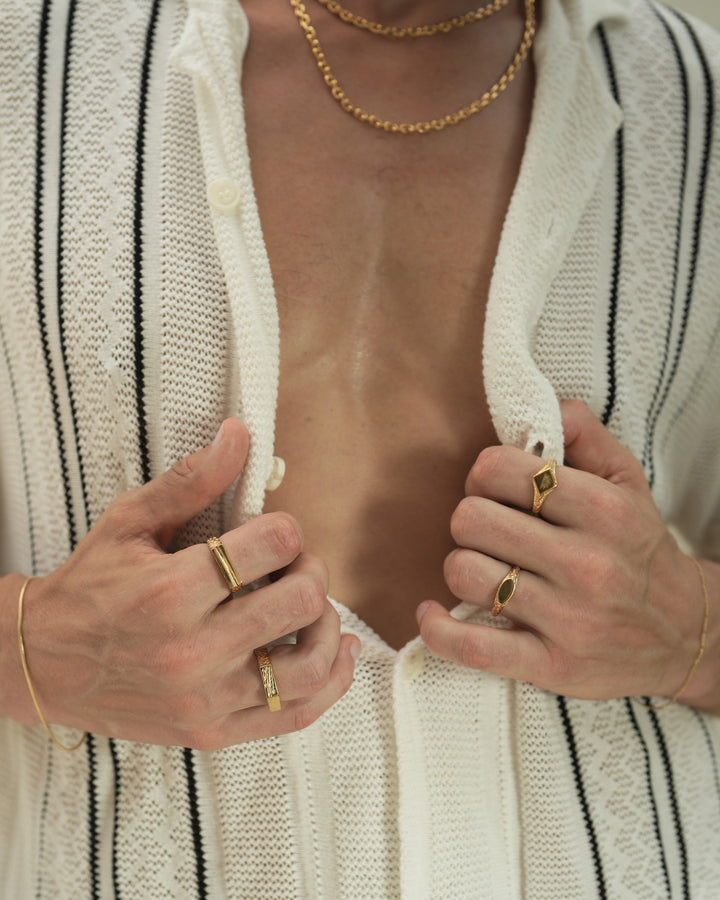 Founder of Nusands Corey Micari front view zoomed in on chest wearing 18k gold layered shackled necklaces, hands both grasping the inside of button up shirt at chest hieght. Right hand wearing roped in bracelet, line in the sand and 6 base rings, left hand wearing signature chain, suns out and mirrored signet rings.
